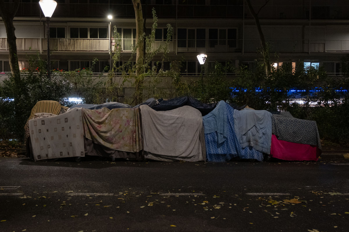 Sleeping bag and personal belongings on a Paris pavement at night, below a sign reading 'Caresse'