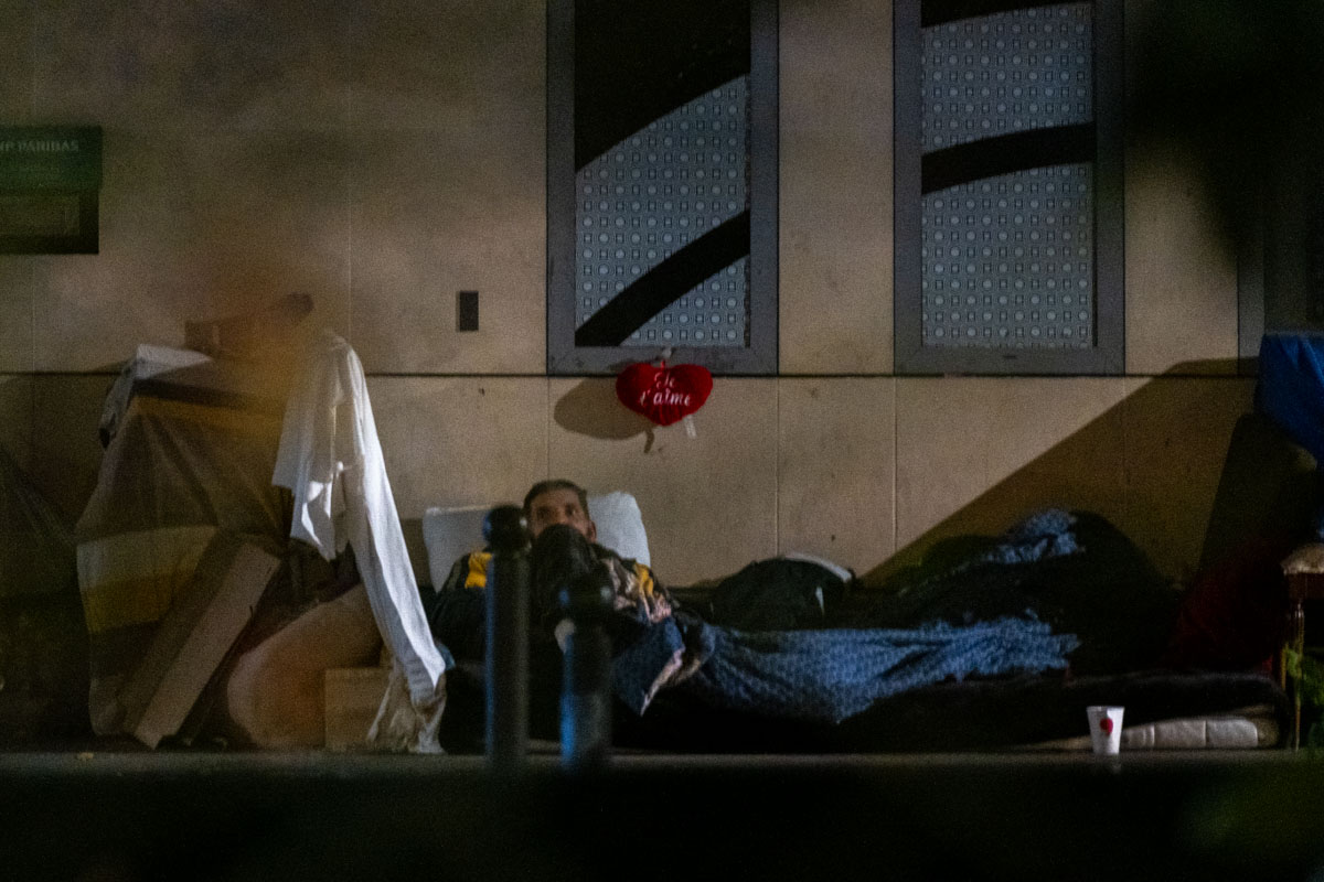 Person sheltering under an umbrella beside a mattress on a wet Paris street at night