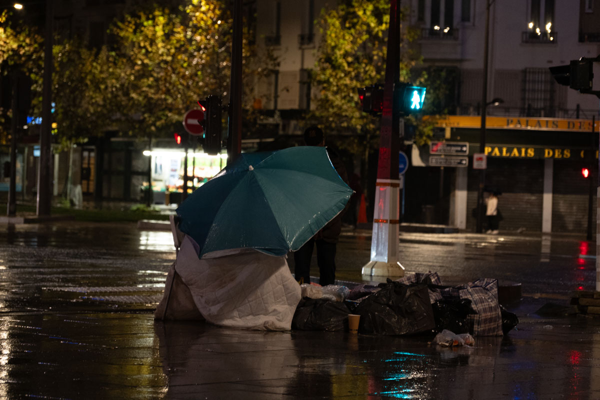 Homeless man lying on a mattress at night in Paris, with a red heart balloon reading 'Je t'aime' on the wall above