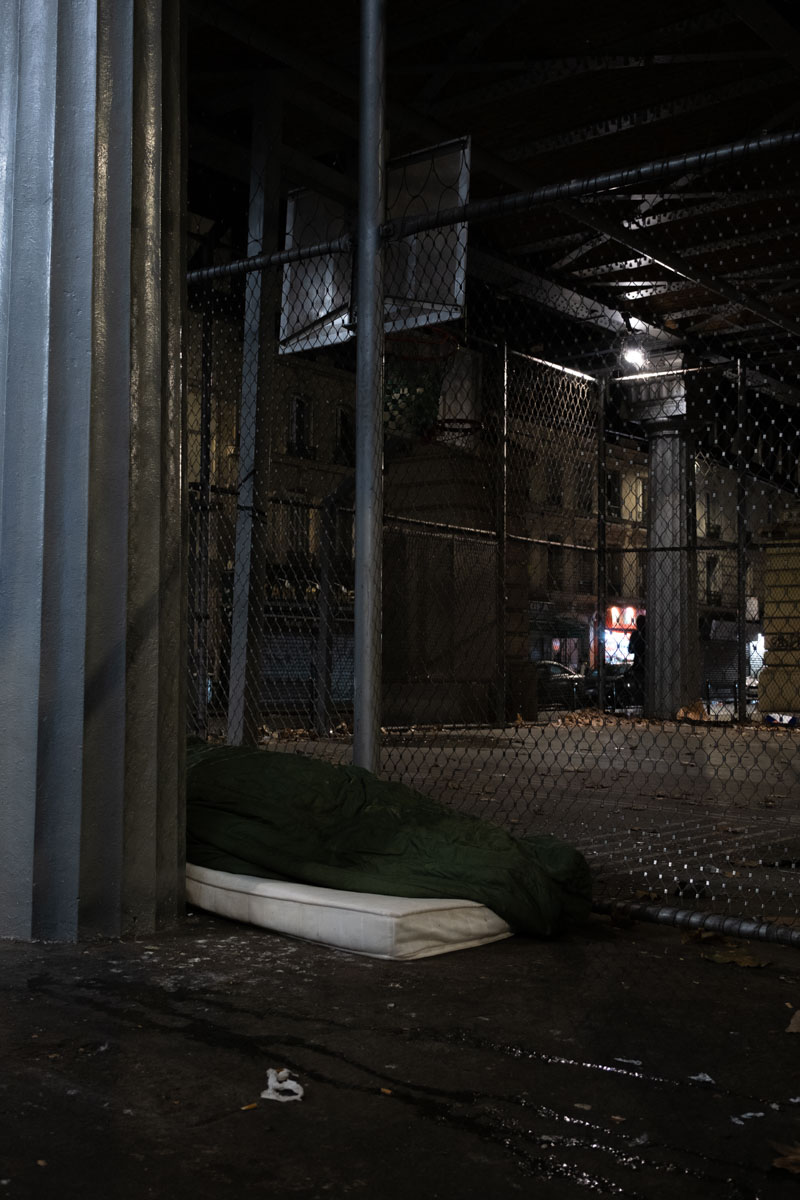 Group of homeless people sleeping under a Paris metro viaduct at night, near a lit brasserie
