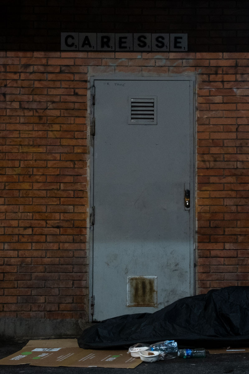 Row of mattresses and blankets forming a makeshift encampment along a Paris pavement at night